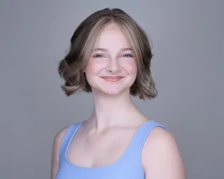 Young actor with wavy bob hairstyle smiling warmly in light blue tank top against gray backdrop